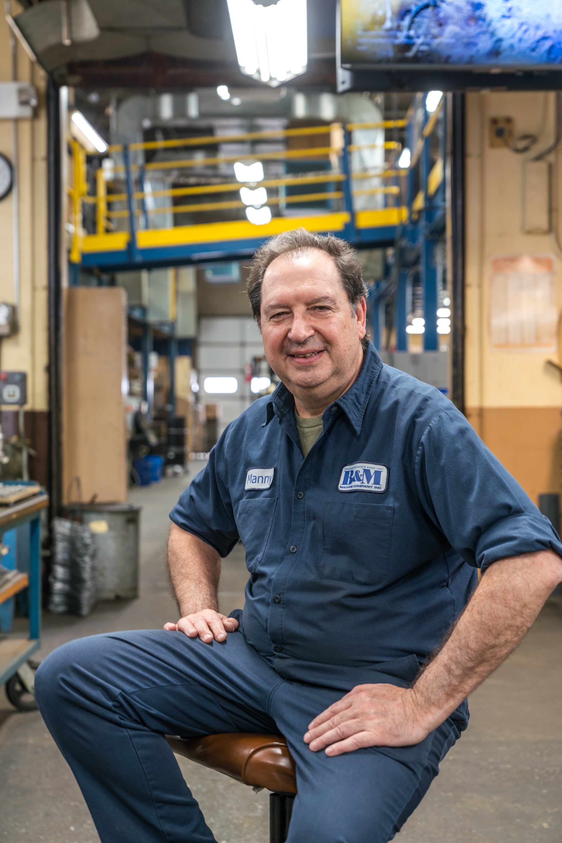 Man in a blue mechanic's uniform sitting on a stool in a workshop.